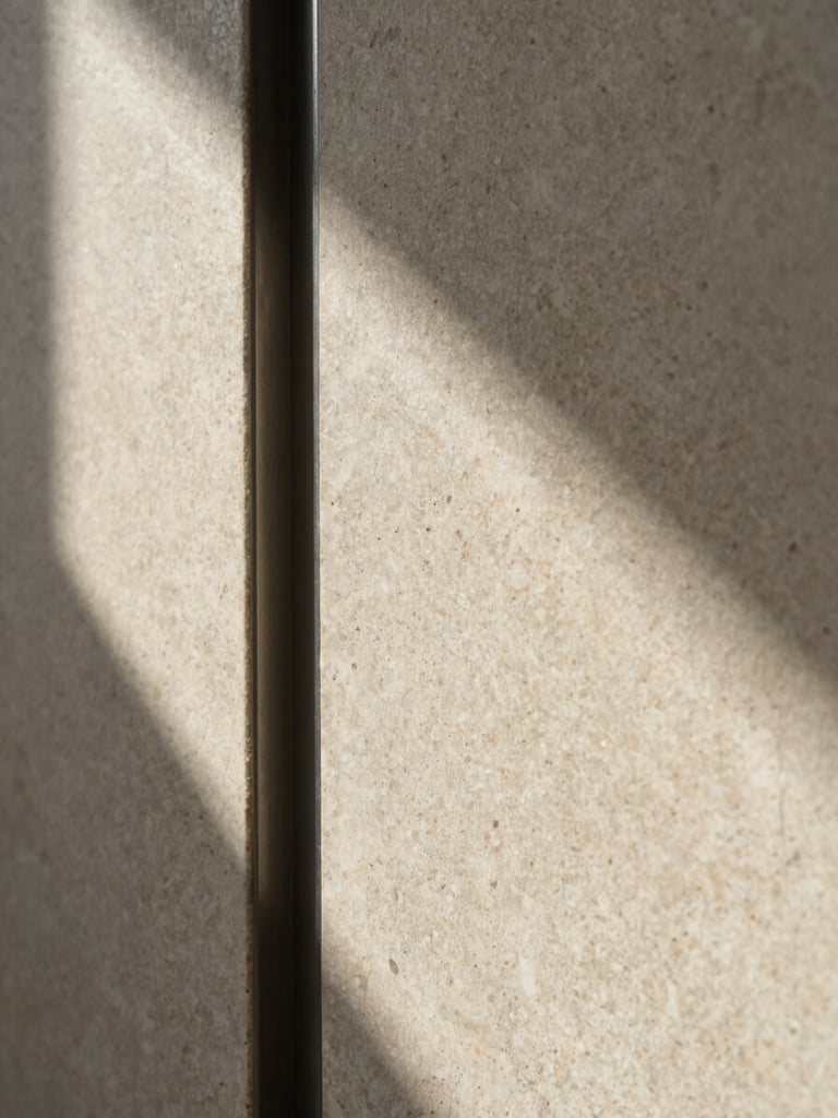 Extreme close-up of aged travertine wall panel meeting a dark steel reveal strip, raking sidelight from the left casting sharp shadows across the stone surface texture, empty architectural interior, no people