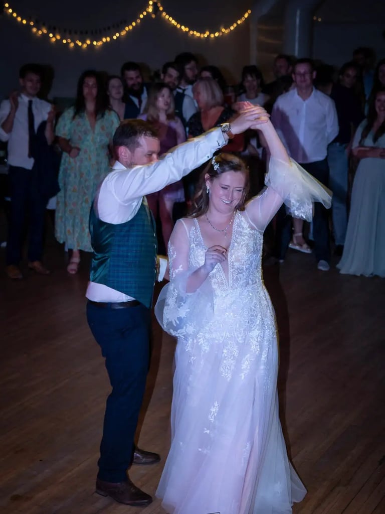 A smiling bride in a lace gown and groom in a green vest dance their first dance at a wedding reception.
