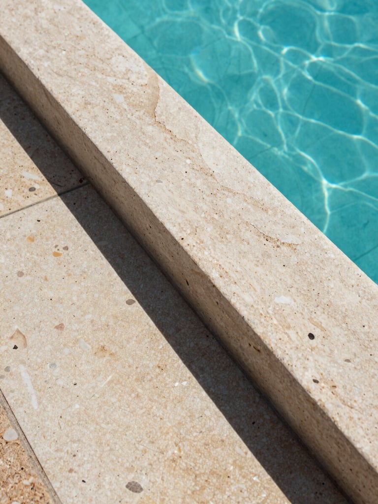 Close-up of natural stone coping and pool edge detail, gresite tile texture visible beneath clear water, Mediterranean midday light casting sharp shadows, documentary framing, no people, architectural precision