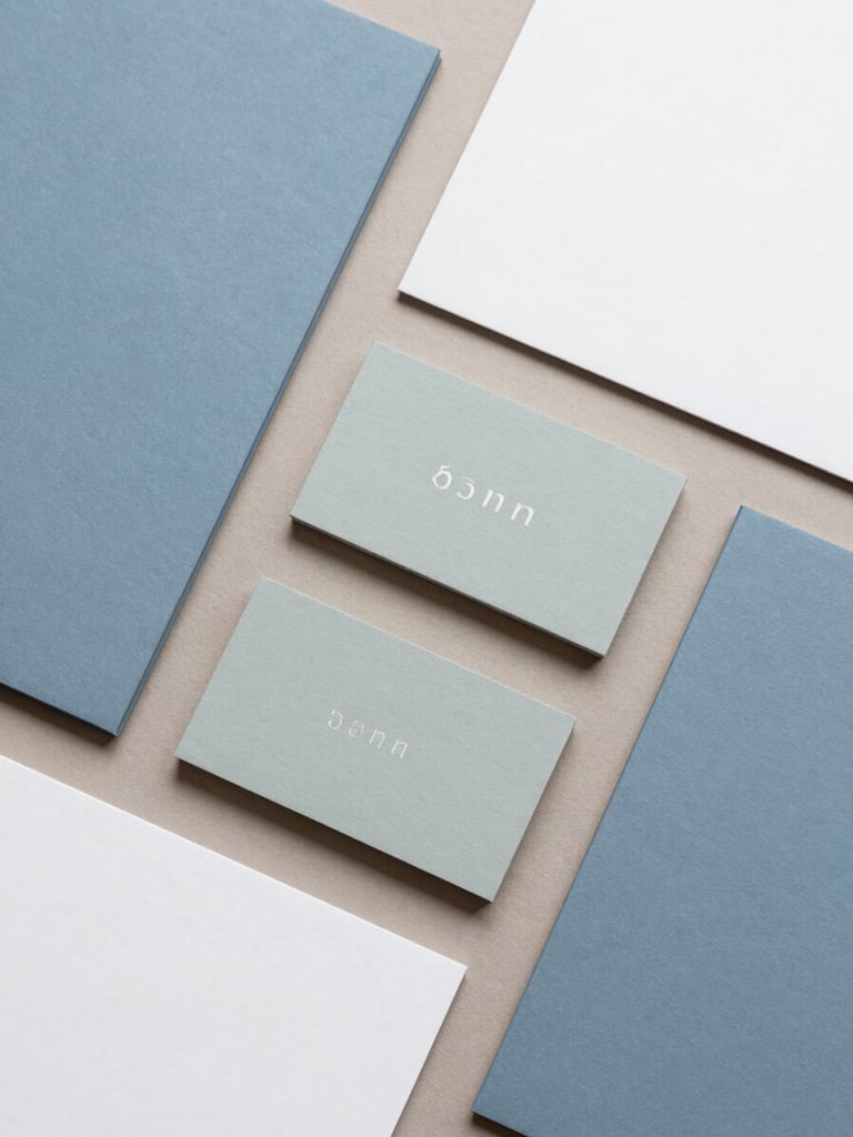 Top-down flat lay of coordinated brand collateral, soft slate blue and muted sage business cards, clean stationery aligned at 90-degree angles on a warm taupe surface, soft diffused overcast daylight, 35mm