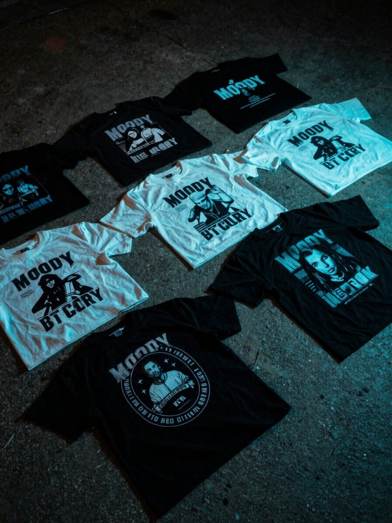 Moody flat-lay of graphic streetwear t-shirts on a rough concrete surface, dramatic shadows, low-key lighting with subtle cyan highlights, showing high-contrast print details.