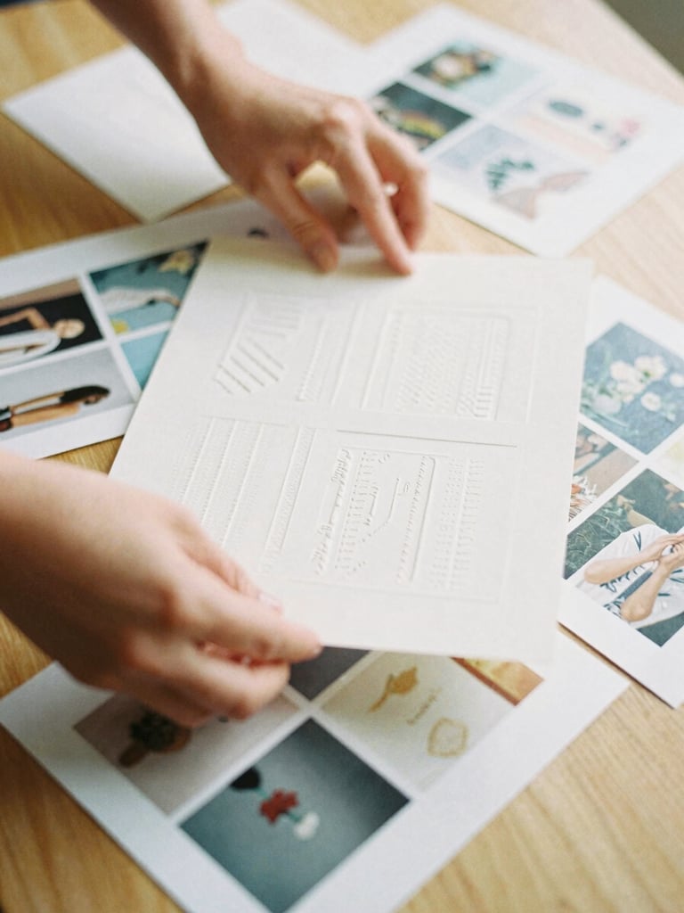 Close-up of a designer's hands arranging tactile paper layout samples and editorial photographs on a light oak table, soft side-lighting, warm tones.