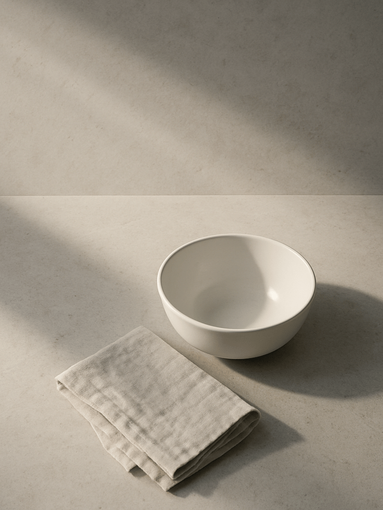 Close-up overhead composition of a minimalist kitchen counter — a single white ceramic bowl, a folded linen cloth, and cool morning light raking across pale stone surface, vast negative space above