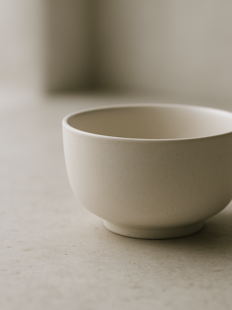 Close-up of a carefully arranged minimalist surface — a single clean ceramic object resting on a pale stone countertop, soft diffused daylight from the left, shallow depth of field, emphasis on texture and negative space, no people