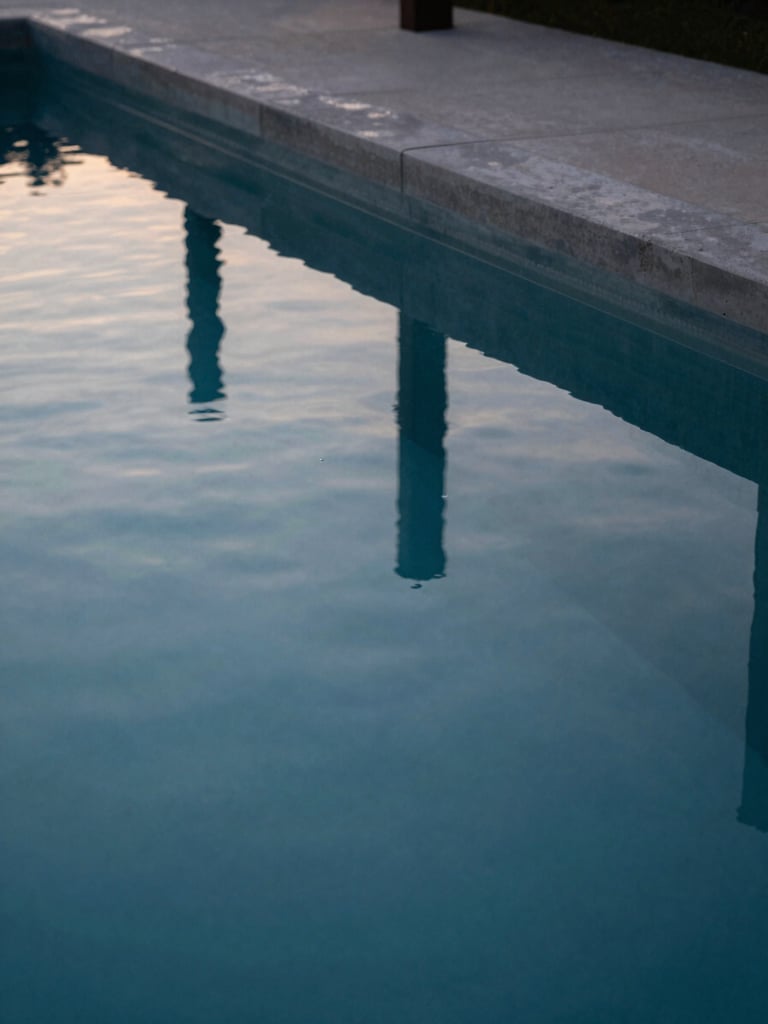 Extreme close-up of still, mirror-like water surface reflecting clean concrete lines of an architectural estate pool at twilight, minimal composition.