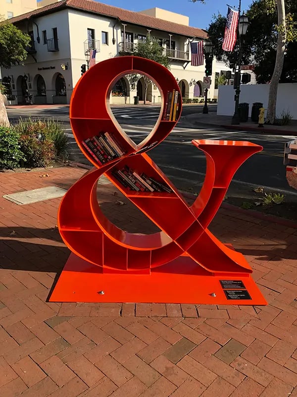 An innovative book-sharing box designed as a large red ampersand, encouraging community reading on a city street.