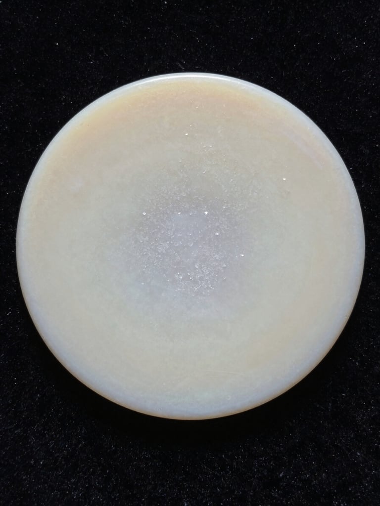 Extreme close-up of a pale wax disc resting on black velvet, soft directional light from the left revealing crystalline surface texture, no labels, no hands, no face — purely material