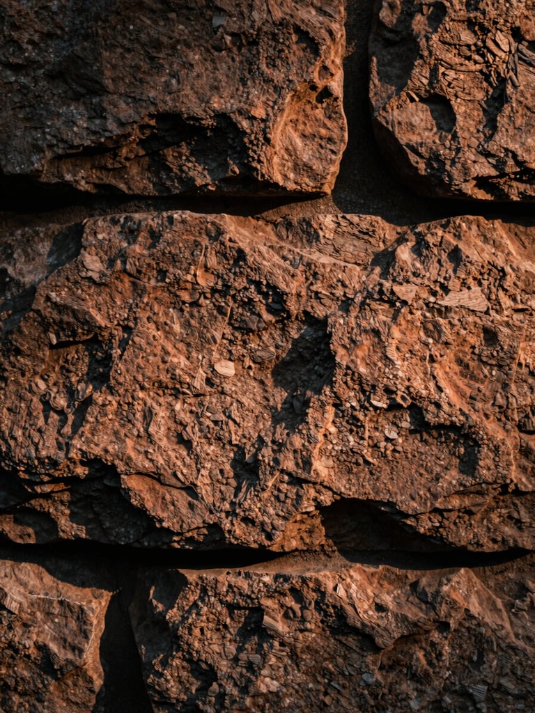 Extreme close-up of a rough stone wall surface, warm raking side-light casting deep shadows across the texture, terracotta and charcoal tones, no people, tactile and still, art-film editorial quality
