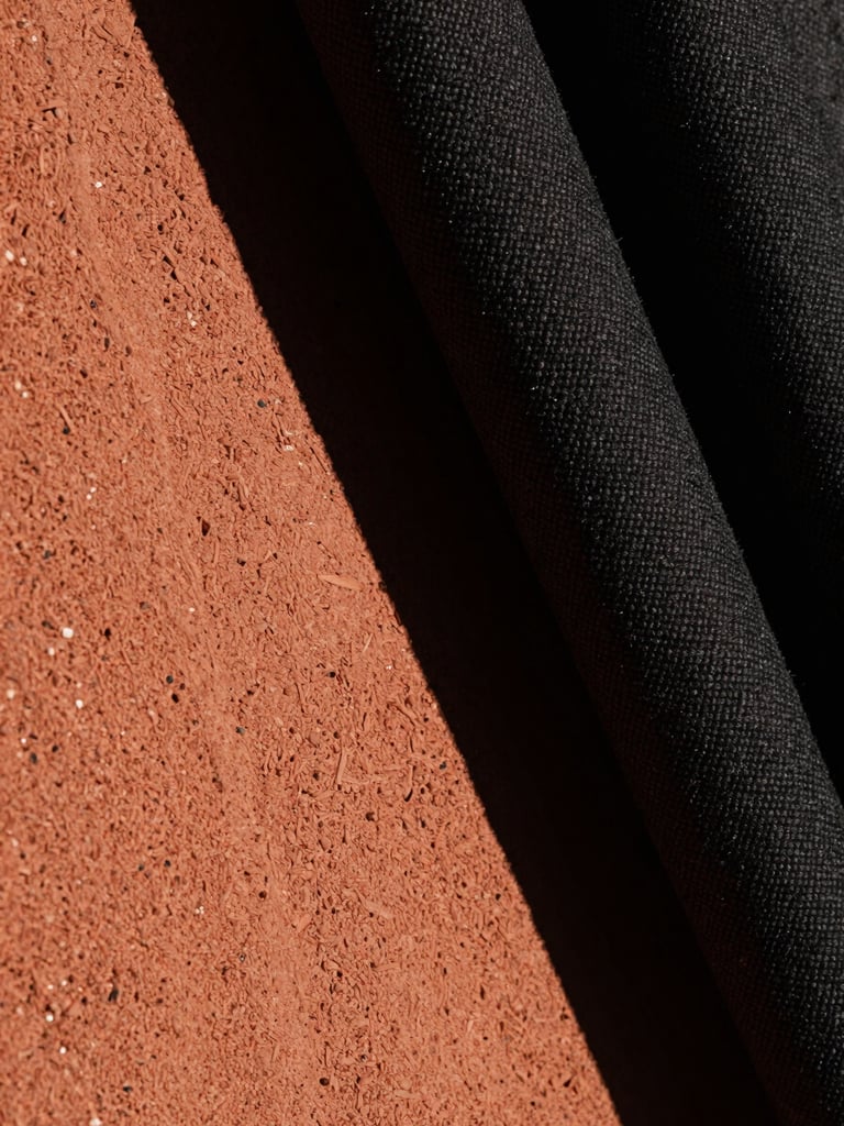 Extreme close-up portrait of raw, textured concrete grain meeting heavy, dark-woven textile fabric. High-contrast chiaroscuro lighting with a single warm terracotta light source casting deep, dramatic shadows.