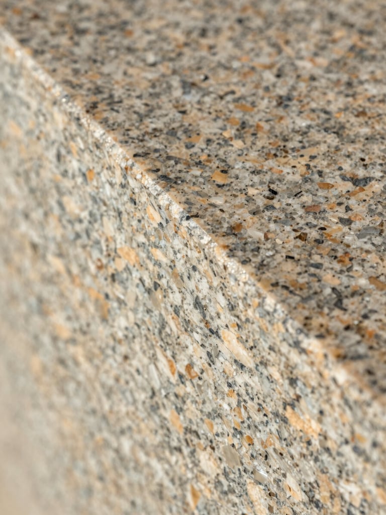 Extreme close-up overhead of a polished granite slab edge profile, directional daylight raking across the surface to reveal crystal grain and finish texture, warm neutral tones, no color correction, full-frame crop showing material reality