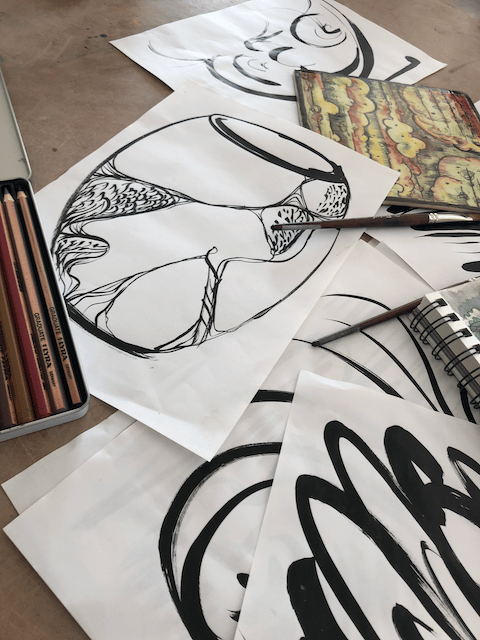 A display of black ink drawings with a vase sketch on top, pencils and notebook