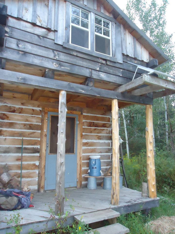 the porch of a rustic wood cabin, with a cooler for water resting on a makeshift shelf