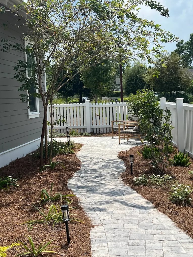 Curving stone paver walkway in a backyard garden with pine straw mulch and a white picket fence.