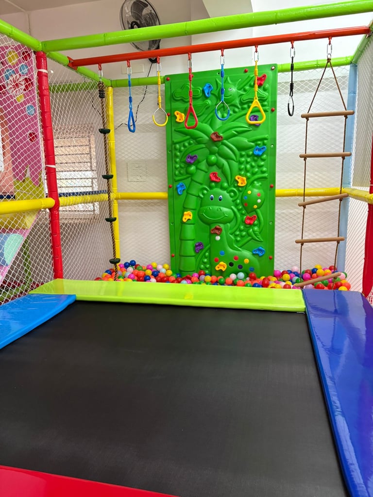 Indoor playground featuring a trampoline, ball pit, climbing wall, and hanging gym rings for kids.