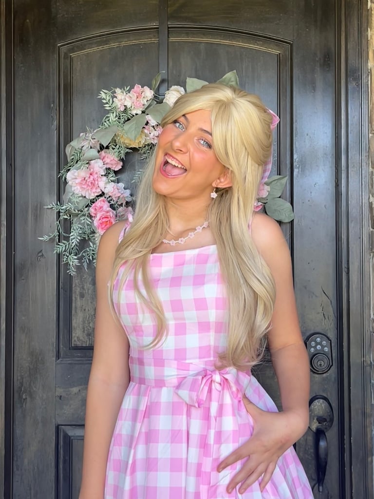 A smiling woman wearing a blonde wig and pink gingham dress in front of a front door floral wreath.