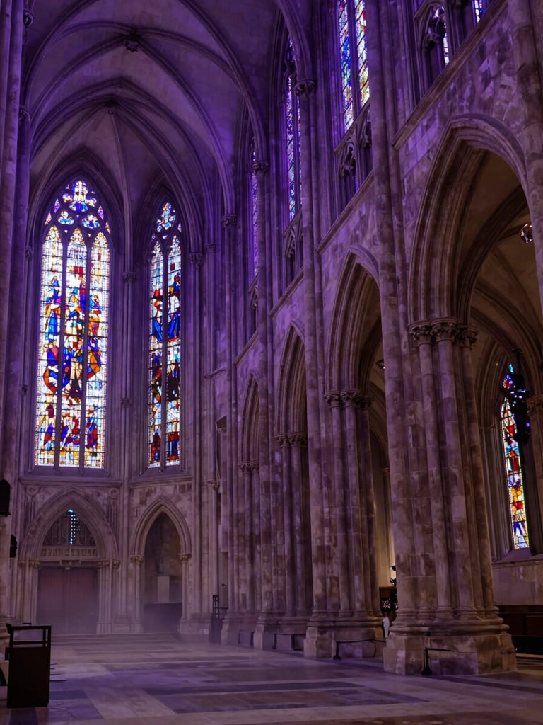 Wide low-angle view of gothic cathedral nave interior, deep violet ambient light filtering through stained glass windows far left, stone vaulted arches receding into shadow on the right, mist pooling along the floor, solemnly cinematic