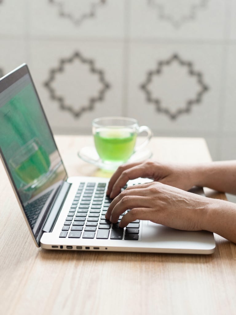Close up of hands typing on a modern laptop on a clean wooden desk, a cup of green tea nearby, soft warm morning light, minimalist Islamic geometric pattern in the background