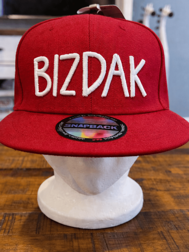 A red snapback hat with white embroidered BIZDAK text displayed on a foam head over a wooden table.