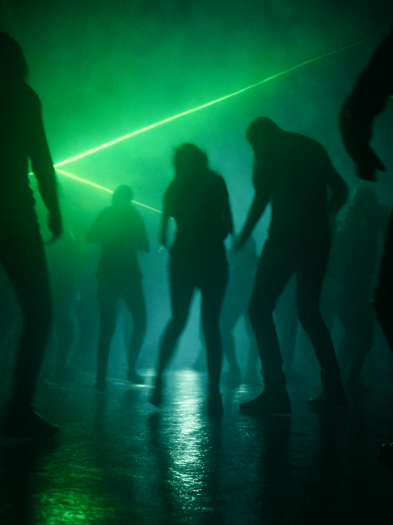 Wide panoramic dancefloor interior of a dark underground club, crowd silhouettes from waist-down moving in unison, twin green laser beams cutting diagonally through heavy smoke from upper left to lower right, floor wet with light reflections — no faces visible, motion blur on the bodies