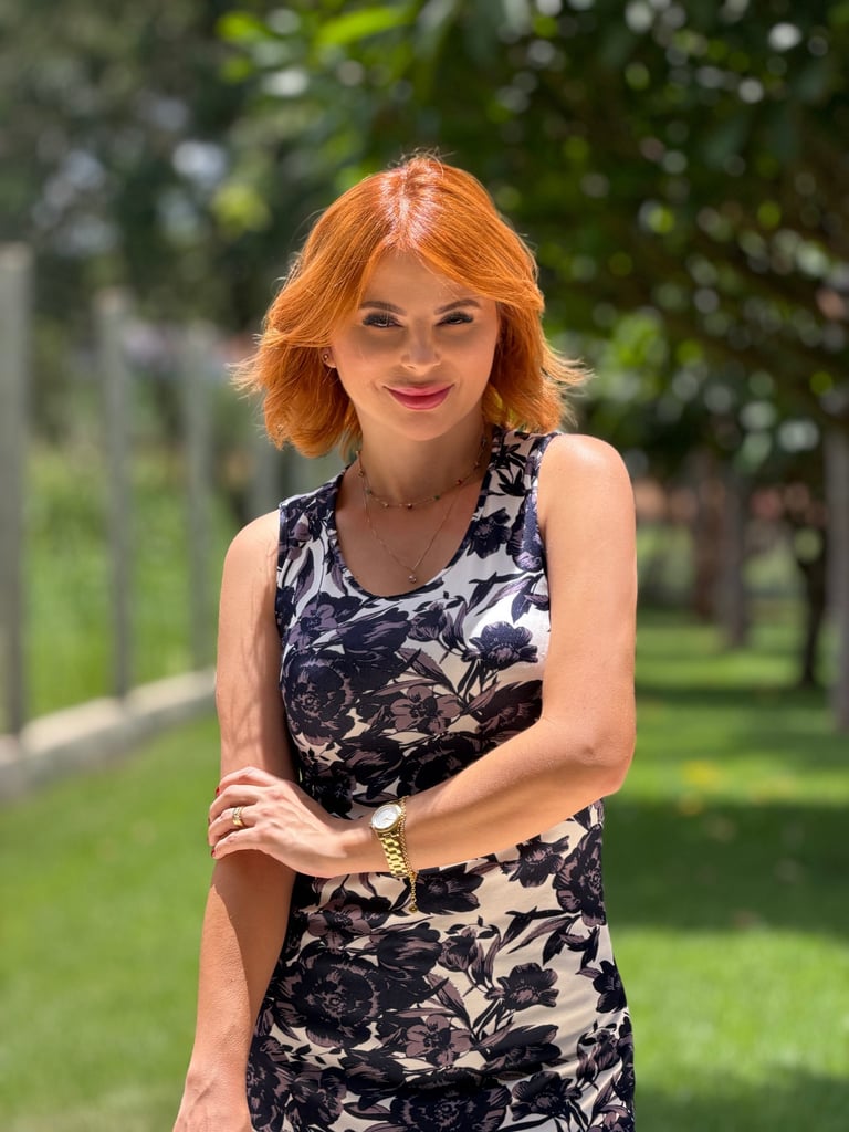 A smiling redhead woman in a floral print sleeveless dress posing outdoors in a sunny park.