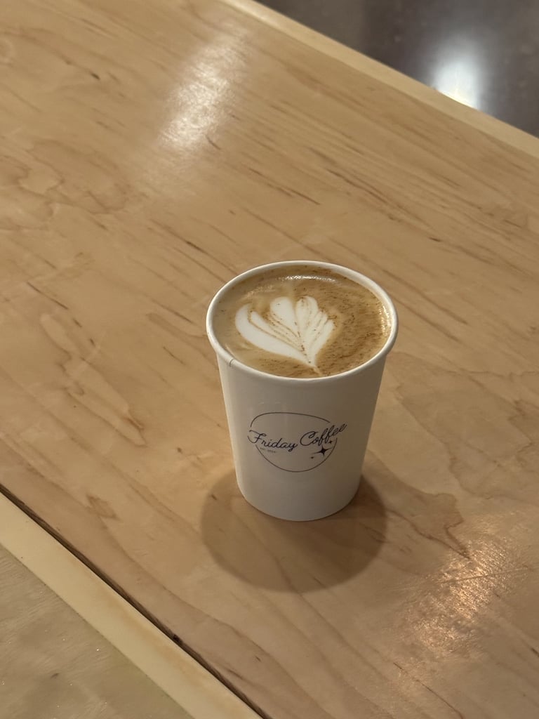 A hot latte with heart-shaped foam art in a white Friday Coffee paper cup on a wooden table.