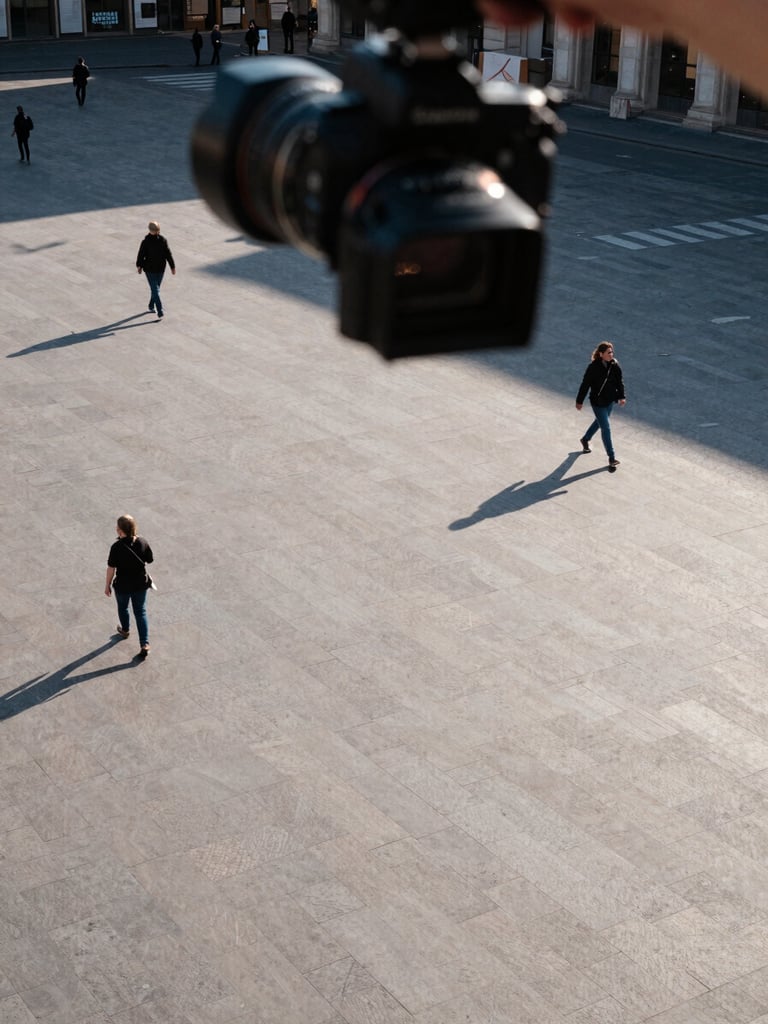 Corner-angle overhead shot looking down from a camera mount at a wide European public square, late afternoon daylight casting long precise shadows across paving stones, a few pedestrians mid-stride visible from above, the camera housing visible in the upper foreground as the compositional anchor, cool natural light, no faces identifiable