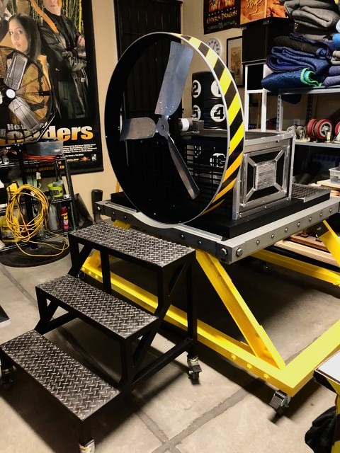 A magician's illusion prop featuring a large industrial fan on a yellow and black striped platform with metal steps, ready
