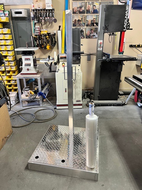 Industrial aluminum diamond plate pallet wrapper stand with a roll of clear stretch film in a workshop.
