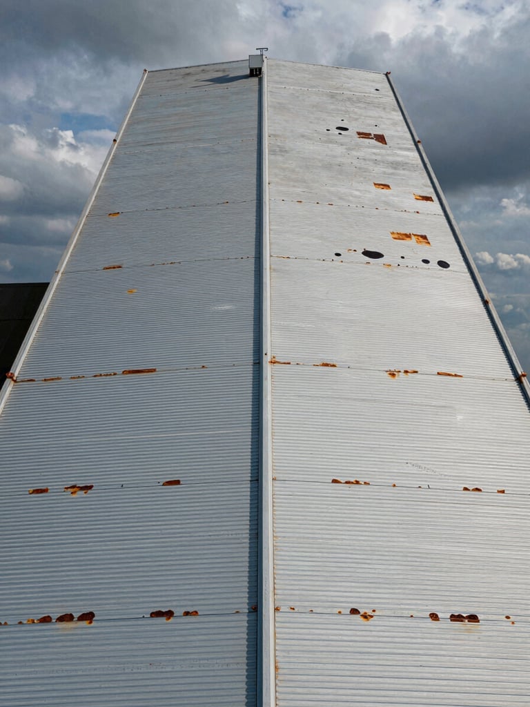 Wide aerial drone perspective looking straight down at the full span of a large industrial warehouse roof, corrugated steel panels visible with rust patches and ponding marks, overcast industrial sky at edges, drone shadow faintly visible on the surface, no people