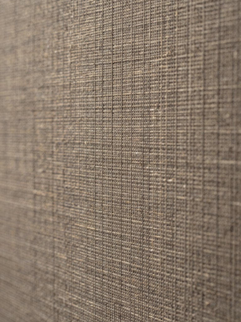 Extreme close-up of a fabric acoustic panel surface, woven textile weave texture in warm gray-tan under flat cool north-facing light, tight macro framing revealing material depth and fiber structure, no people