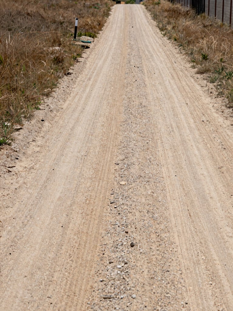 Low ground-level approach shot along a compacted dirt road leading into an undeveloped parcel, natural daylight from overhead, dry grass on both sides, a visible boundary marker post in the mid-ground, neighboring fence line at the right edge, no people, no structures