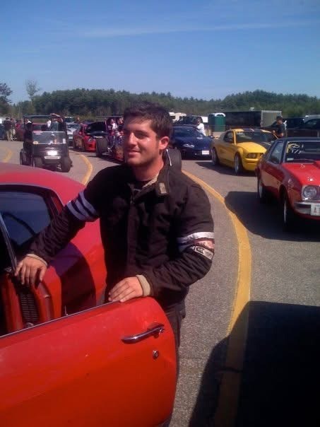 Don abenante racer in a black jacket stands by a red muscle car at a busy drag racing track.