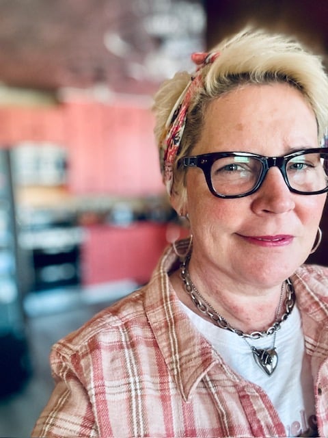 White woman in chunky black glasses and a plaid shirt, wearing a bandana in her hair.