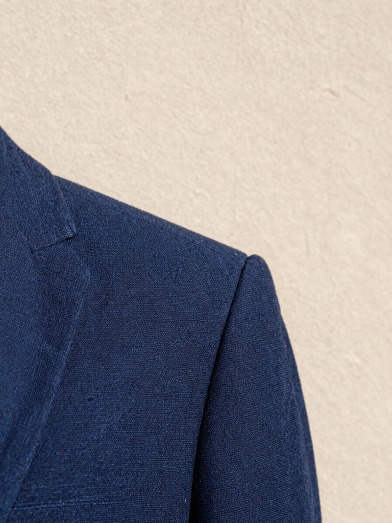 Close portrait crop, natural north-facing daylight, a single folded garment of deep indigo woven fabric resting against a pale clay-toned wall, fine weave texture visible in sharp focus, generous negative space above, no hands or props