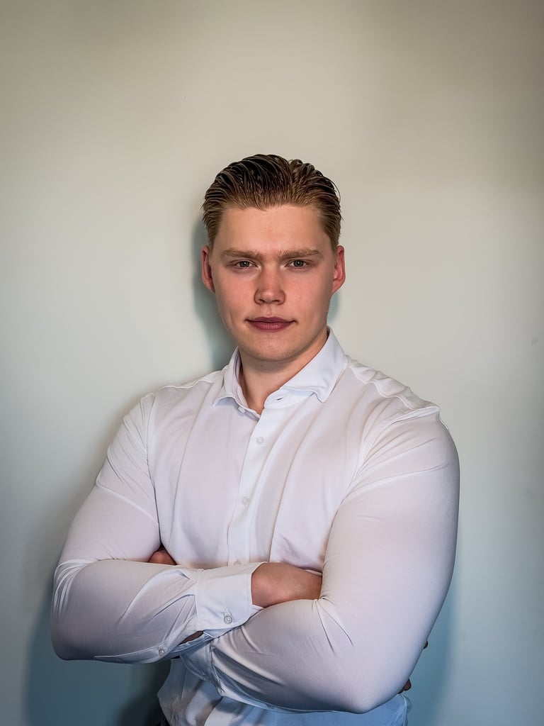 Professional portrait of a young man with crossed arms wearing a white dress shirt against a neutral wall.