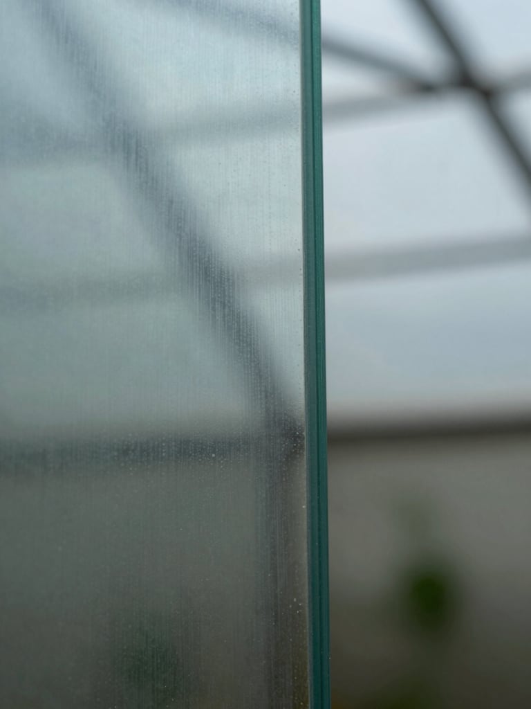Extreme close-up of tempered greenhouse glass panel edge, natural north-facing daylight raking across the surface revealing internal structure and edge clarity, macro framing, no glare, cool diffuse light