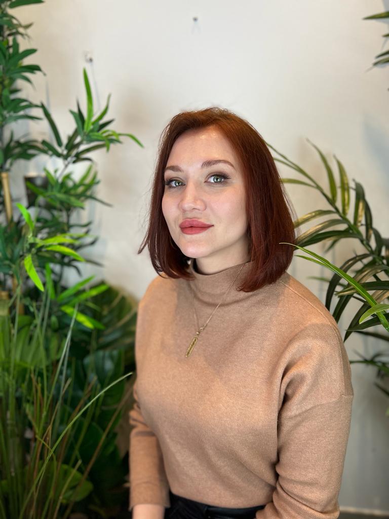 Woman with a professional auburn bob haircut wearing a tan turtleneck sweater among green plants.