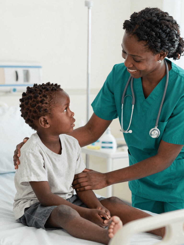 A smiling female pediatric nurse in green scrubs comforting a young boy sitting on a hospital bed.