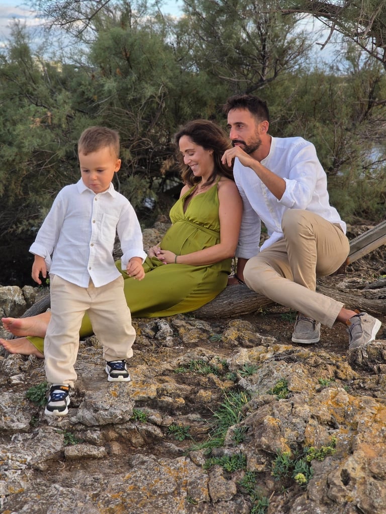 A family of three sitting on rocky terrain near a river with greenery in the background, enjoying an outdoor moment.