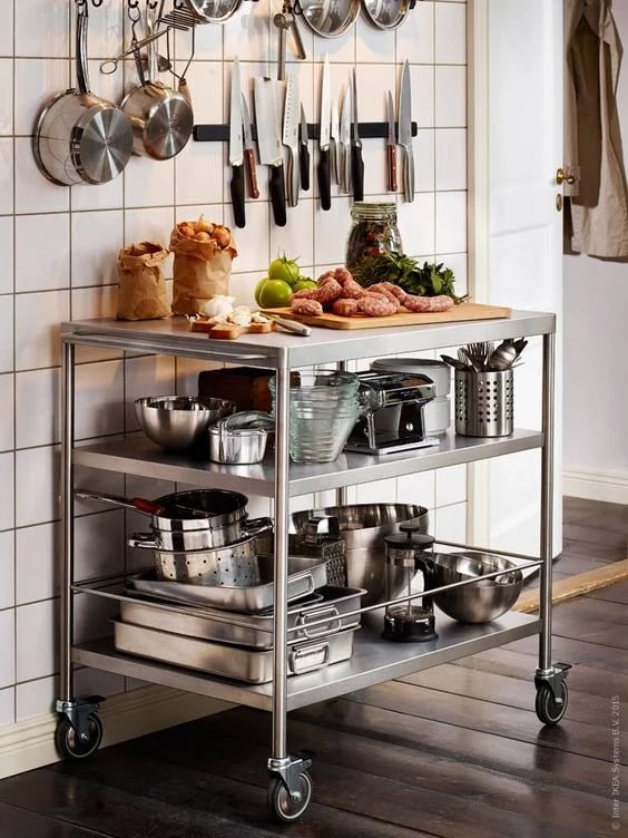 A mobile stainless steel table with three shelves used for food prep and storing kitchenware against a tiled wall.