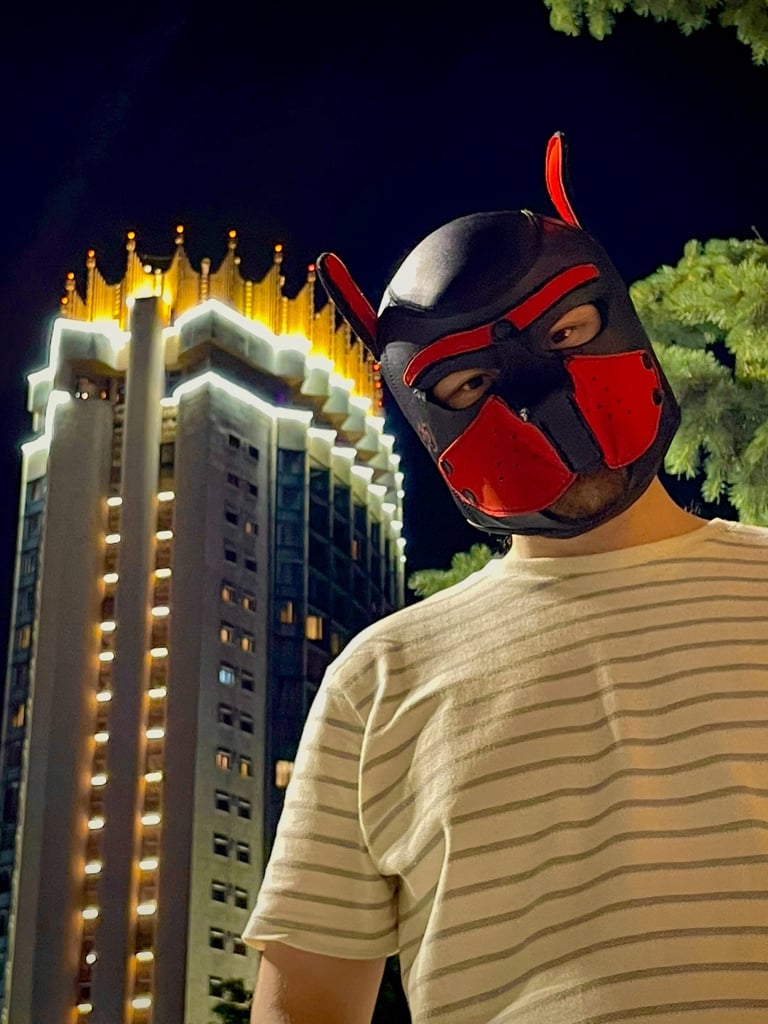 Young man in a pup mask, posing by a tower at night.