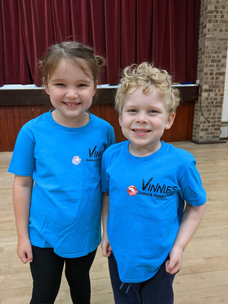 Two smiling children in blue Vinnies Dance & Theatre School t-shirts on a wooden dance floor.