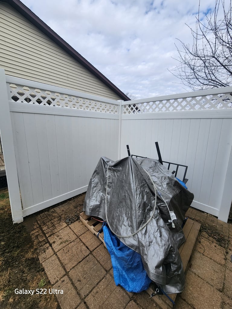 Backyard patio equipment protected by a heavy-duty silver tarp inside a white vinyl privacy fence.