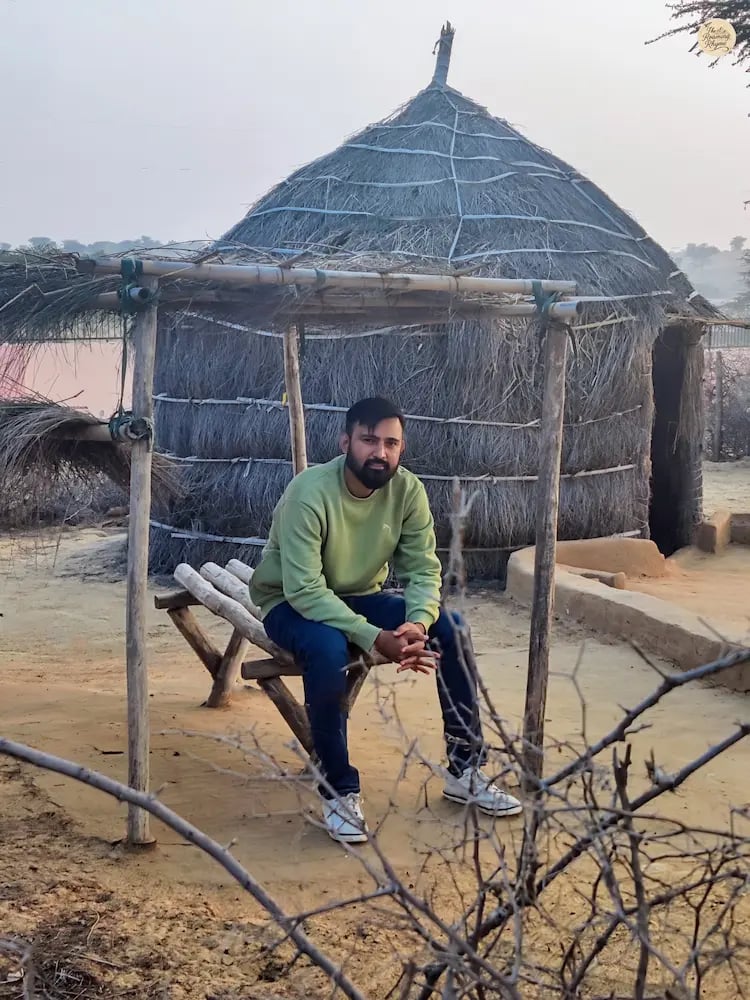 Person sitting by a traditional Rajasthani hut in Raisar dunes, Bikaner, experiencing desert calm.