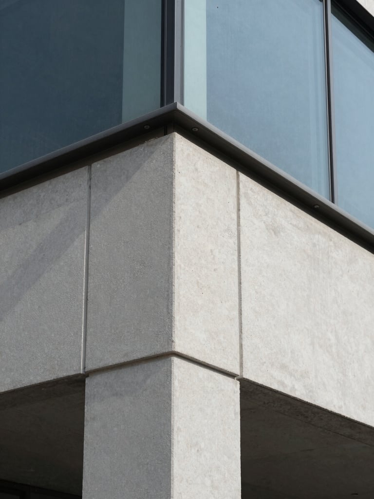 Close-up architectural detail shot, right-edge bleed, of a facade material junction — concrete panel meeting a glazed curtain-wall system, structural fixings visible, daylight raking across the surface revealing texture and tolerances, no people, documentary framing