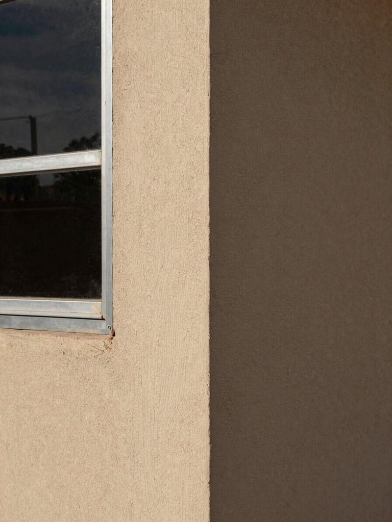 Portrait-format close detail of a freshly rendered exterior wall corner meeting a metal window frame, Grande Comore, natural afternoon light, visible texture of local sand-cement mix, no people