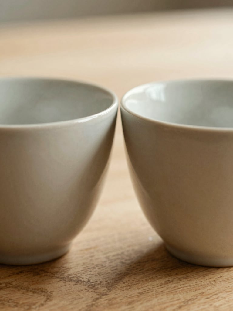 Macro shot of two ceramic cups standing close together on a textured wooden table, soft morning light, warm beige tones, shallow depth of field
