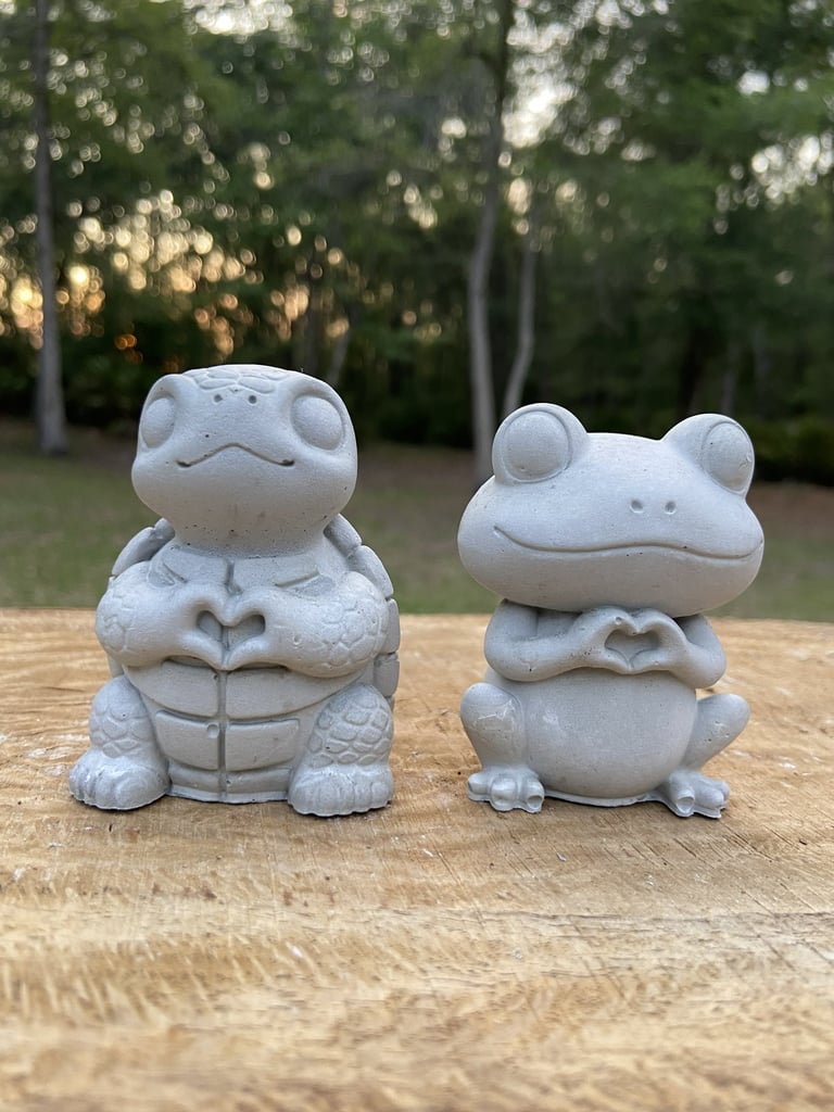 Concrete turtle and frog garden statues making heart shapes with their hands on a wooden surface.