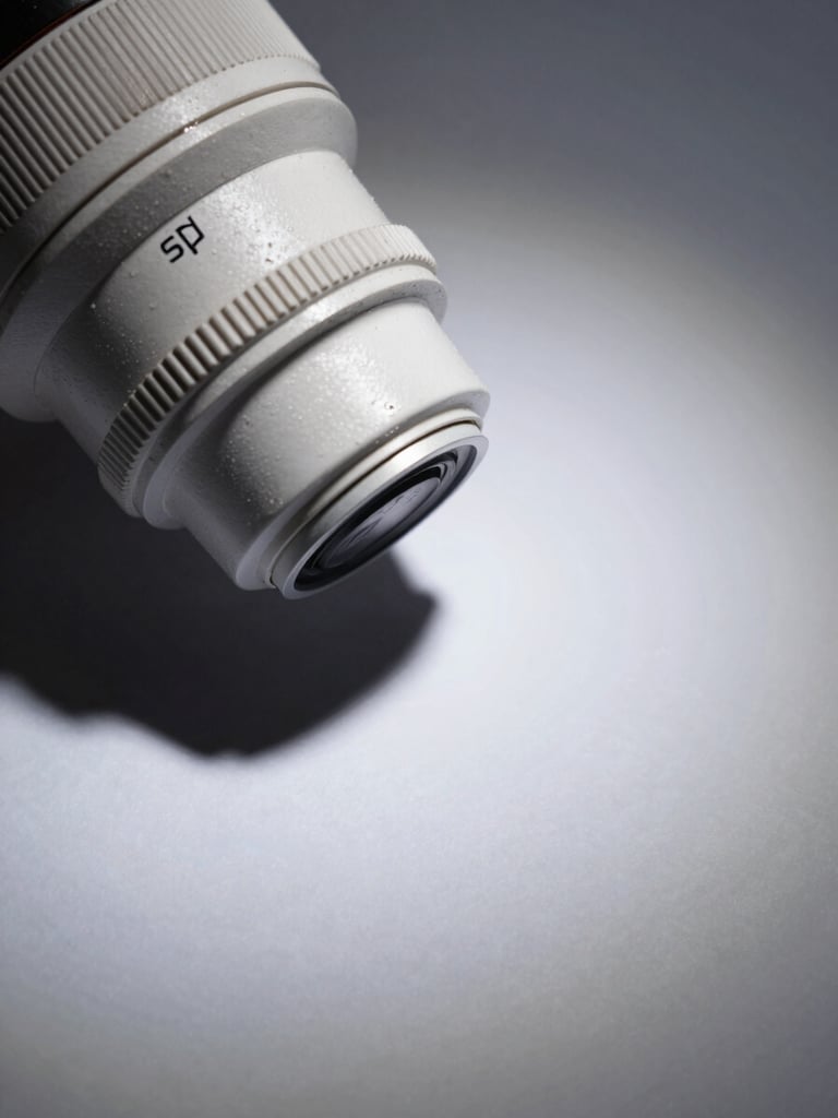 Wide editorial still — close-up of a luxury product on a matte surface, studio strobe lighting from camera-left casting a precise hard shadow, color-correct cool white ambient fill, tight crop showing material texture and typographic label detail, generous negative space to the right