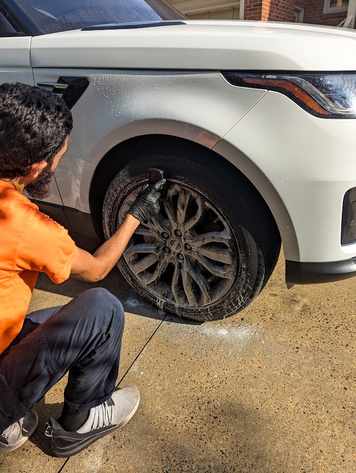 Professional car detailer cleaning a white SUV's alloy wheels with foam soap and a brush.